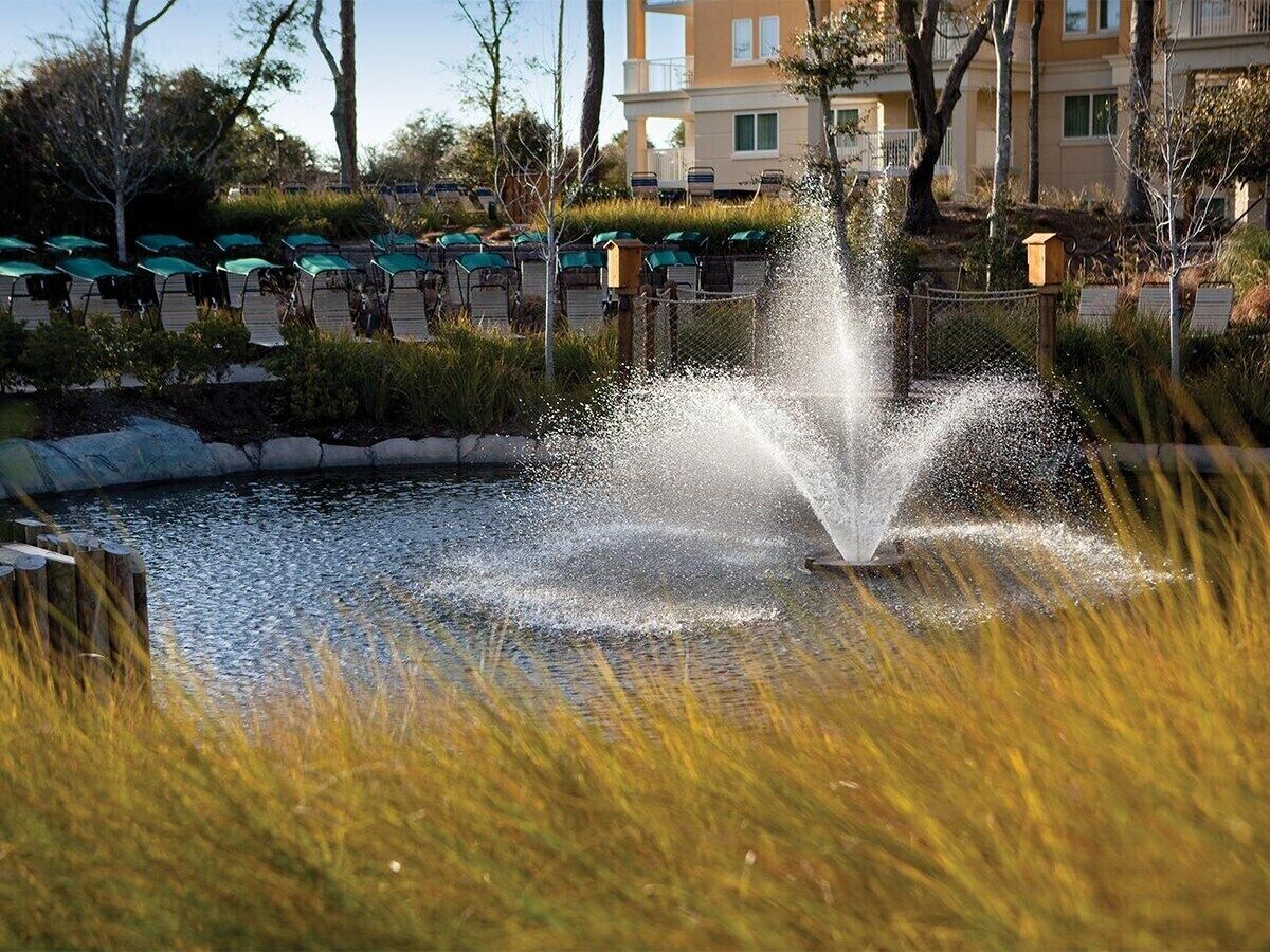 A fountain in a landscaped area with a pool and lounge chairs in the background.