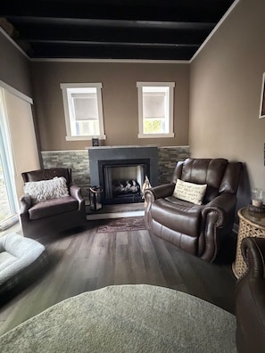 A living room with a fireplace, two windows, leather armchairs, and a patterned rug.