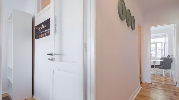Um corredor com uma porta branca, um relógio fixado na parede e uma área de jantar com mesa e cadeiras.
