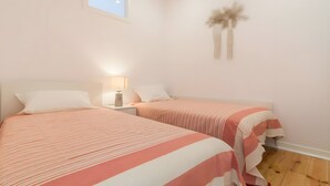 Quarto com duas camas, mesa de cabeceira com abajur, piso de madeira e janela.