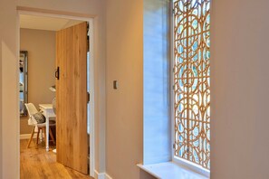 Un pasillo con una puerta de madera que da a una habitación, una ventana con un celosía decorativa y un sillón con un cojín estampado.