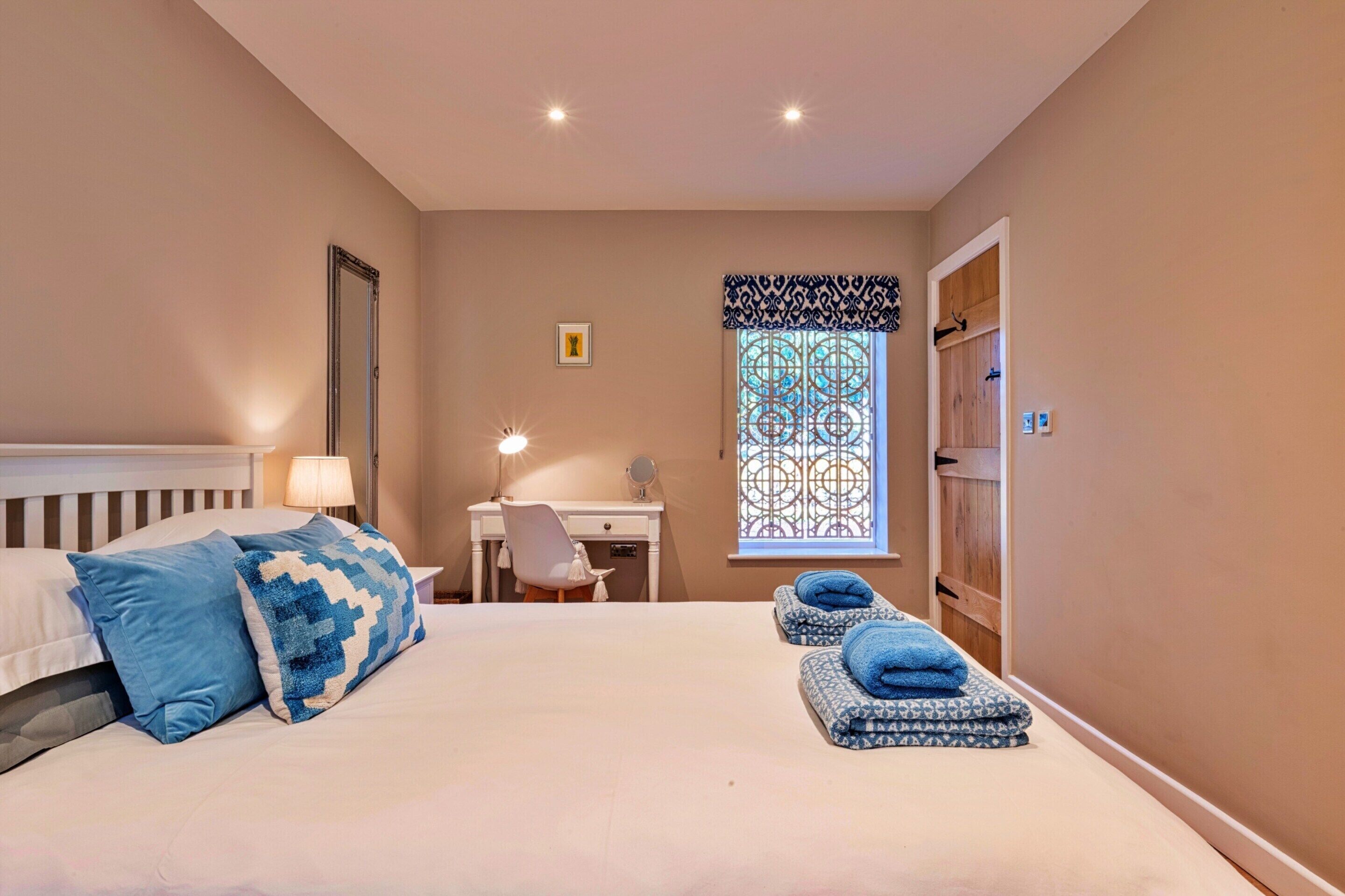 A bedroom with a bed, a desk, a chair, a window with decorative iron bars, and a door.