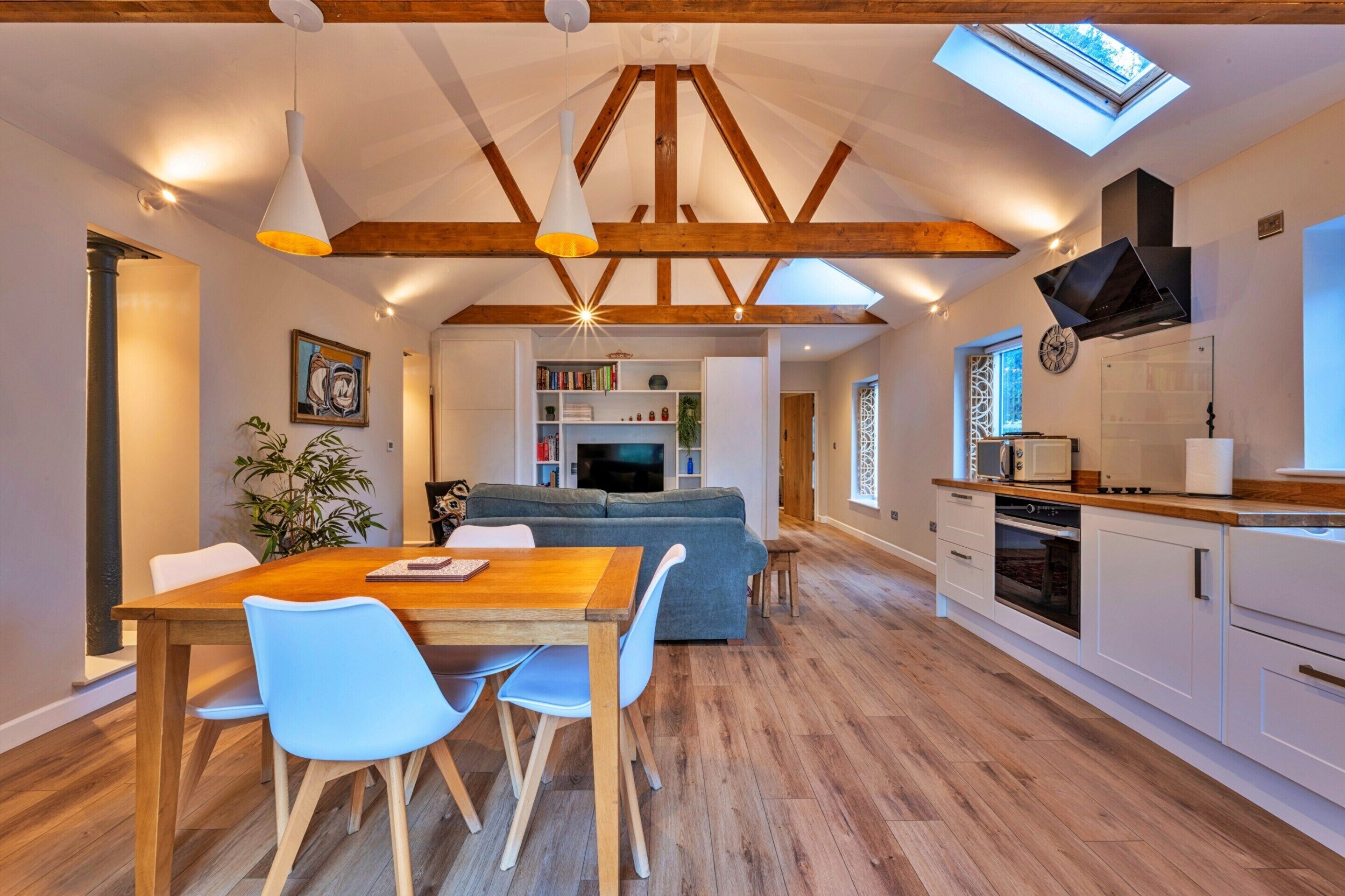 Una cocina moderna con vigas de madera, un comedor con mesa y sillas, una sala de estar con sofá y un tragaluz.