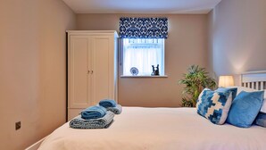 Un dormitorio con cama, ventana con cortina decorativa, puerta, planta en la mesita de noche y lámpara.