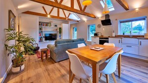 Un interior moderno con vigas de madera a la vista, un comedor con mesa y sillas de madera, una sala de estar con sofá y una cocina con horno empotrado y un ventanal grande.