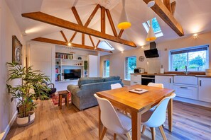 A modern interior with exposed wooden beams, a dining area with a wooden table and chairs, a living room with a sofa, and a kitchen with a built-in oven and a large window. - Romantic Retreat in Lincolnshire (Sleaford)
