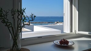 A room with a view of the sea through a large window, a white table with a plate of fruit, and a potted plant. - Parian Pearl · Naousa 180° SeaView · Walk to Beach (Nauosa)