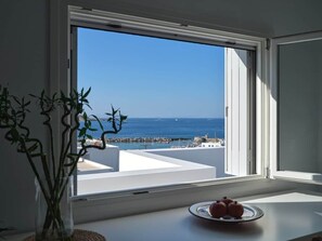 Ein Zimmer mit Meerblick durch ein großes Fenster, ein weißer Tisch mit einer Fruchtschale und ein Blumentopf.