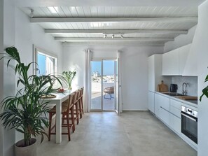 A modern kitchen with white cabinetry, a dining area with wooden chairs, and a large potted plant. - Parian Pearl · Naousa 180° SeaView · Walk to Beach (Nauosa)