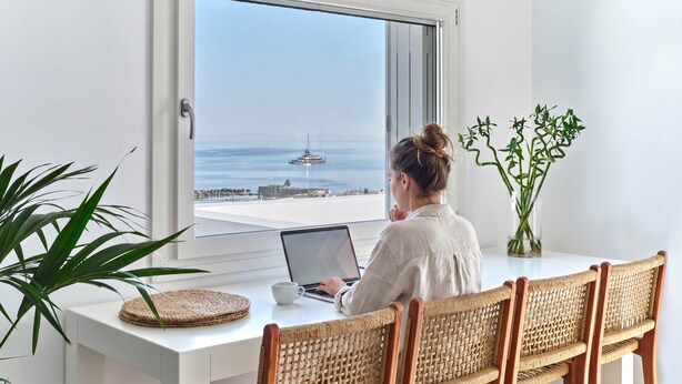A person seated at a white desk with a laptop, facing a large window overlooking a marina with boats. - Parian Pearl · Naousa 180° SeaView · Walk to Beach (Nauosa)