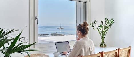 Une personne est assise à un bureau blanc, devant un ordinateur portable, et fait face à une grande fenêtre donnant sur une marina où sont amarrés des bateaux.