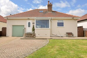 A single-story house with a brick chimney, a green garage door, and a small front yard with a bench. - Beach House With Sea Views (Filey)