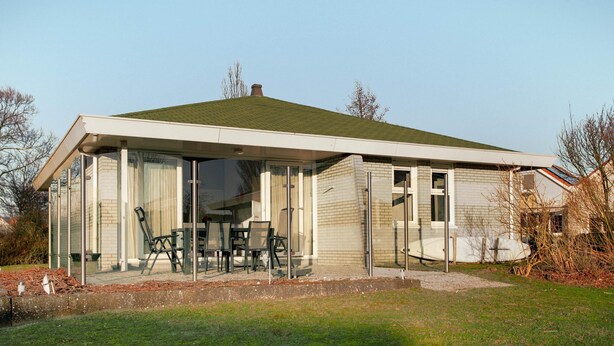 A single-story house with a covered patio area, a dining table with chairs, and a large window. - Detached Waterfront Villa With Garden (Makkum)