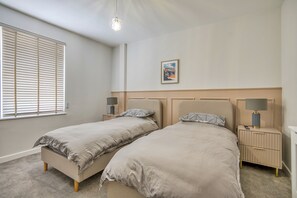 A bedroom with two beds, a nightstand with a lamp, a window with blinds, and a picture on the wall. - Romantic Devon Cottage for 2 (Pembroke Dock)