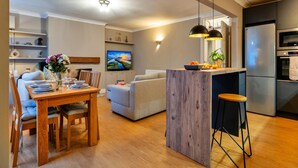 A modern kitchen with a built-in oven and refrigerator, a dining area with a wooden table and chairs, and a living room with a sofa and a TV. - Elegant Apartment in Regency Cheltenham (Cheltenham)