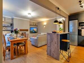 Eine moderne Küche mit Einbauofen und Kühlschrank, ein Essbereich mit Holztisch und Stühlen sowie ein Wohnzimmer mit Sofa und Fernseher.