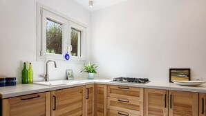 A kitchen with a white countertop, wooden cabinets, a window with a view, a sink, and a stove. - Owl’s Nest · Tsoukalia · Walk to Beach (Τοπική Κοινότητα Αρχιλόχου)