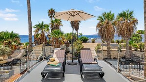 En balkon med to liggestole, en stor parasol og udsigt til havet og palmer.