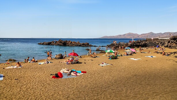 Una playa con gente tomando el sol y nadando, un litoral rocoso y montañas al fondo.