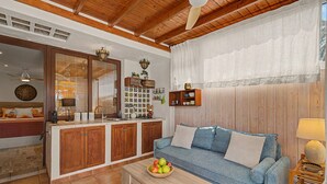 Una sala de estar con techo de madera, un sofá, una mesa de centro con frutas y una zona de cocina con armarios de madera y salpicadero de azulejos.