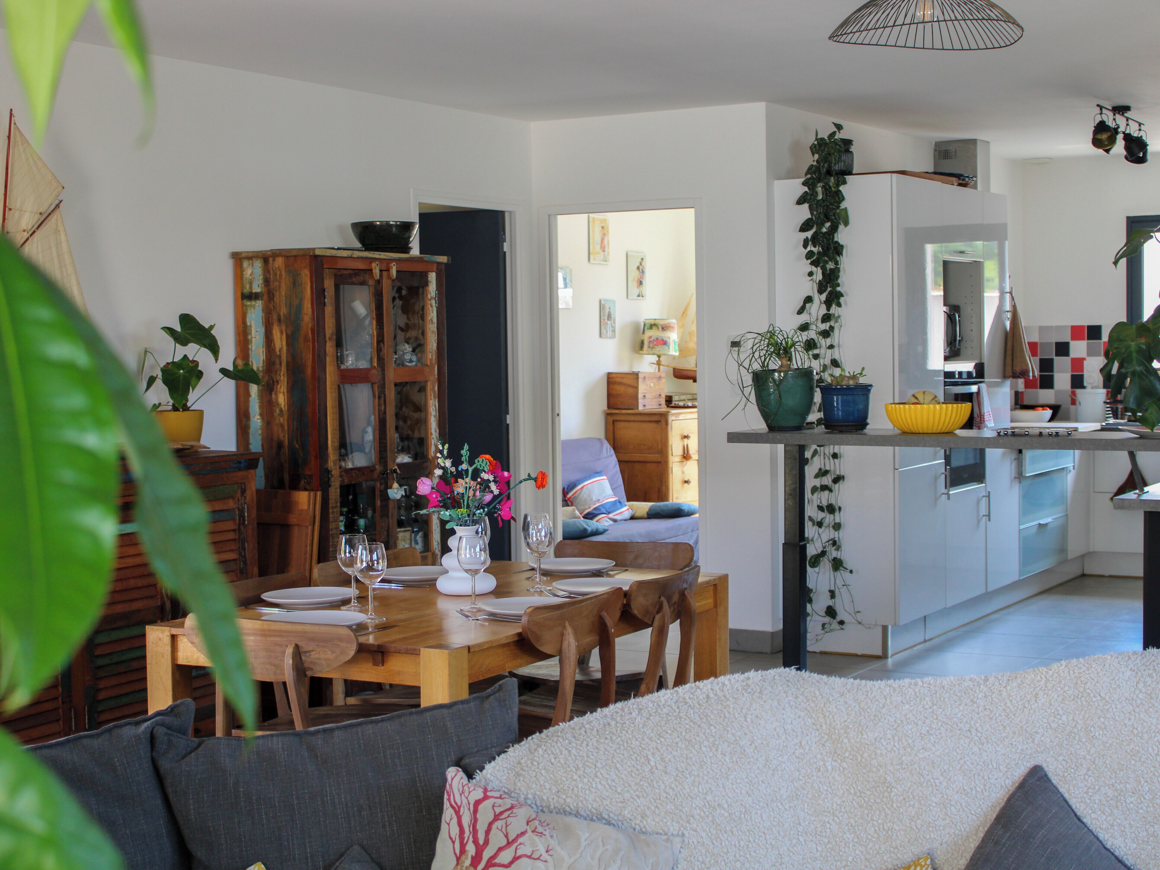 Un coin repas avec une table en bois dressée pour quatre personnes, un canapé avec des coussins et un espace cuisine équipé de placards et agrémenté d’une plante suspendue.
