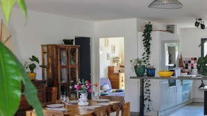 Ein Essbereich mit Holztisch, gedeckt für vier Personen, einer Couch mit Kissen sowie eine Küchenzeile mit Schränken und einer hängenden Pflanze.