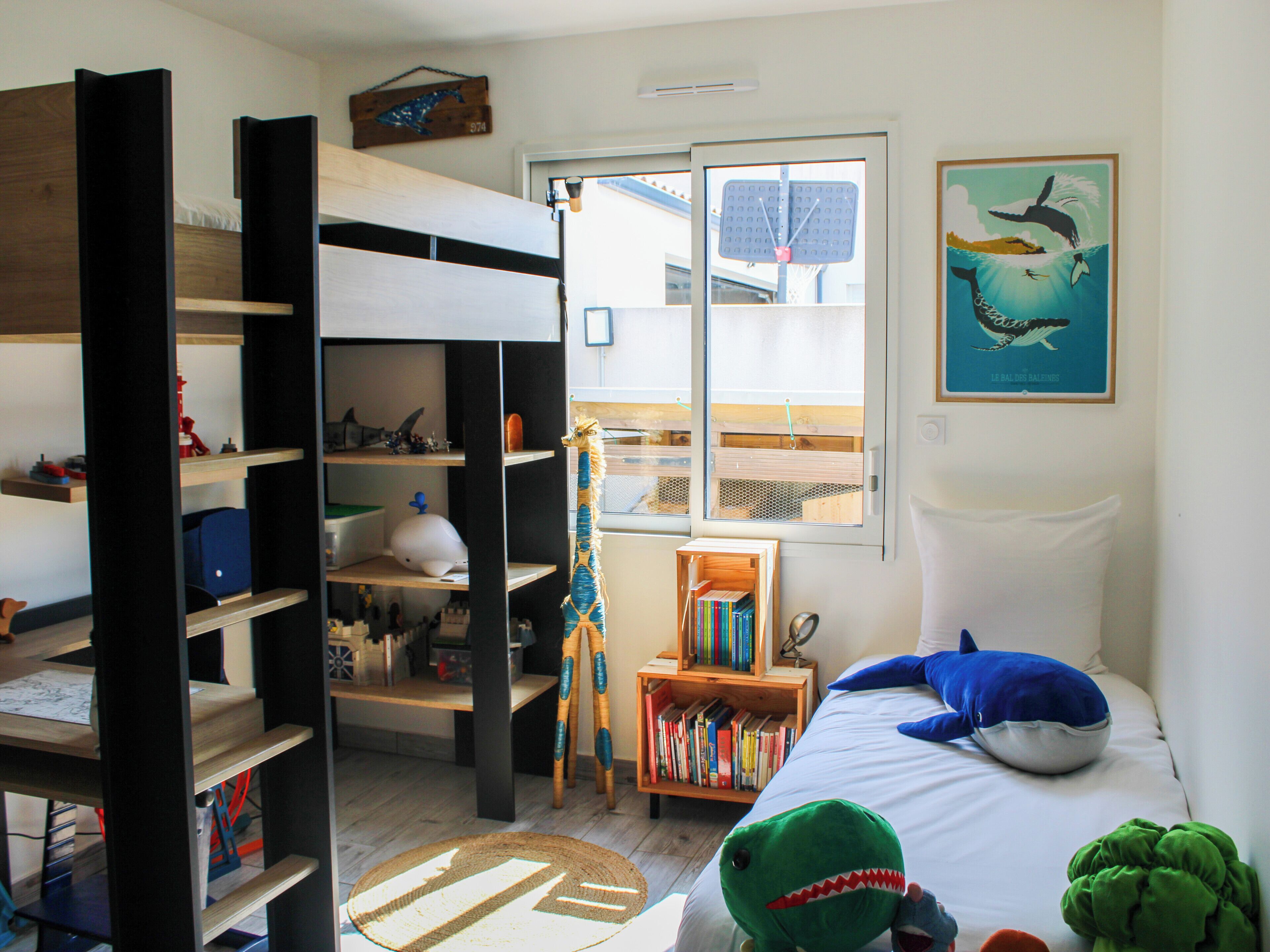 Une chambre d’enfant avec un lit superposé, des étagères remplies de jouets et de livres, une fenêtre donnant sur un paysage agréable et une affiche accrochée au mur.