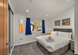 A bedroom with a large bed, a nightstand, a lamp, a window with curtains, and two framed pictures on the wall. - Deluxe Lodge With Hot Tub for 6 (Perth)