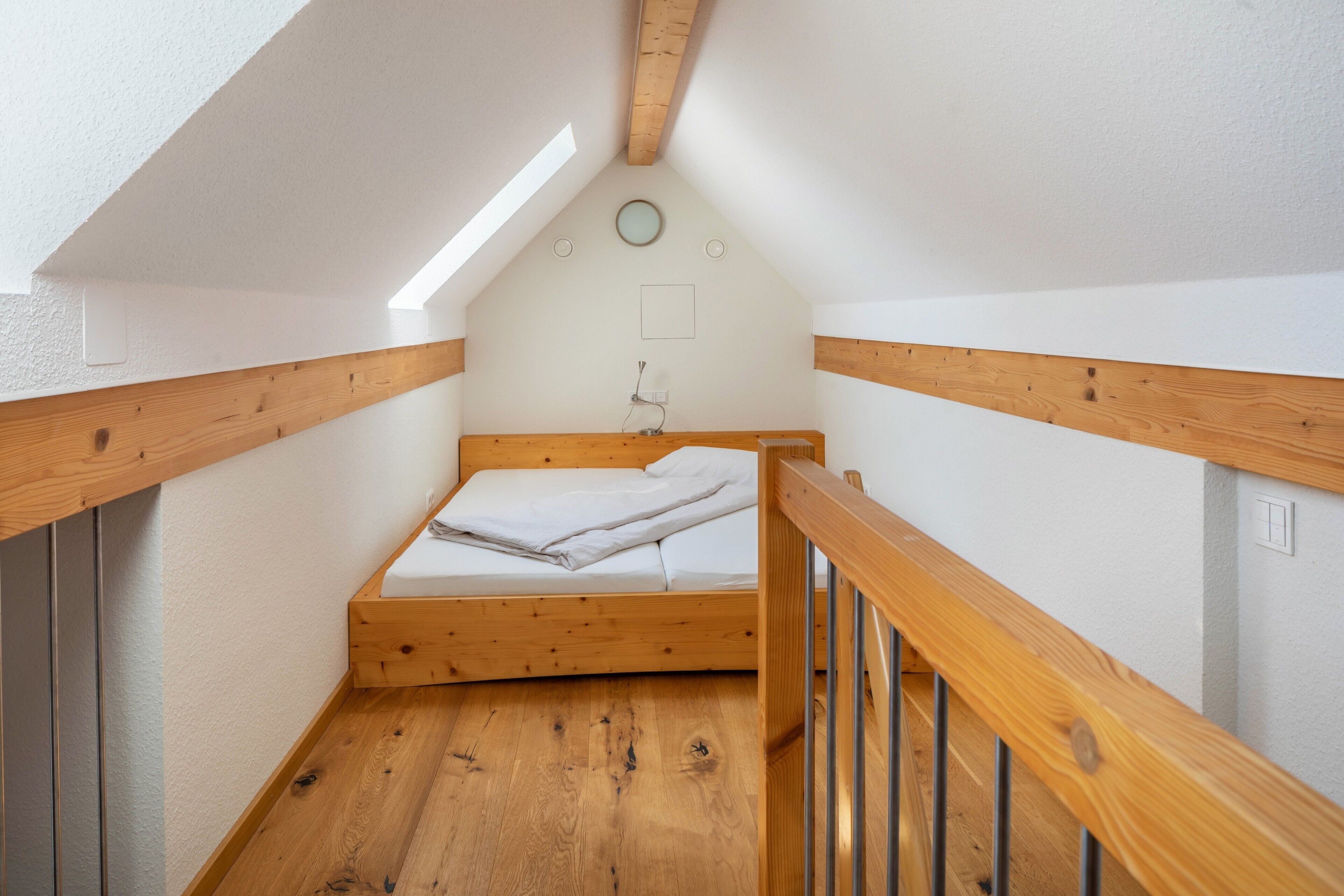Une petite chambre mansardée étroite, avec un plafond en pente, des poutres apparentes, un lit simple et un puits de lumière.