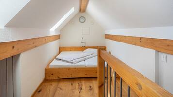 Une petite chambre mansardée étroite, avec un plafond en pente, des poutres apparentes, un lit simple et un puits de lumière.