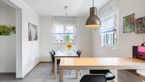 Un espace repas moderne avec une table en bois, des chaises noires et un luminaire suspendu. Il y a deux fenêtres avec des rideaux, et des œuvres d’art sont exposées sur les murs.