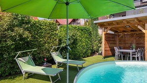Une piscine entourée de verdure et dotée d’une terrasse en bois, avec des chaises longues et un parasol.