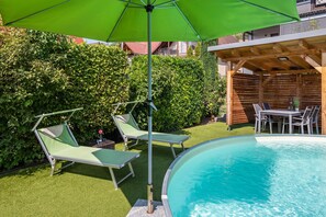 Ein Schwimmbecken mit Liegestühlen und einem Sonnenschirm, umgeben von Grünpflanzen und einer Holzterrasse.