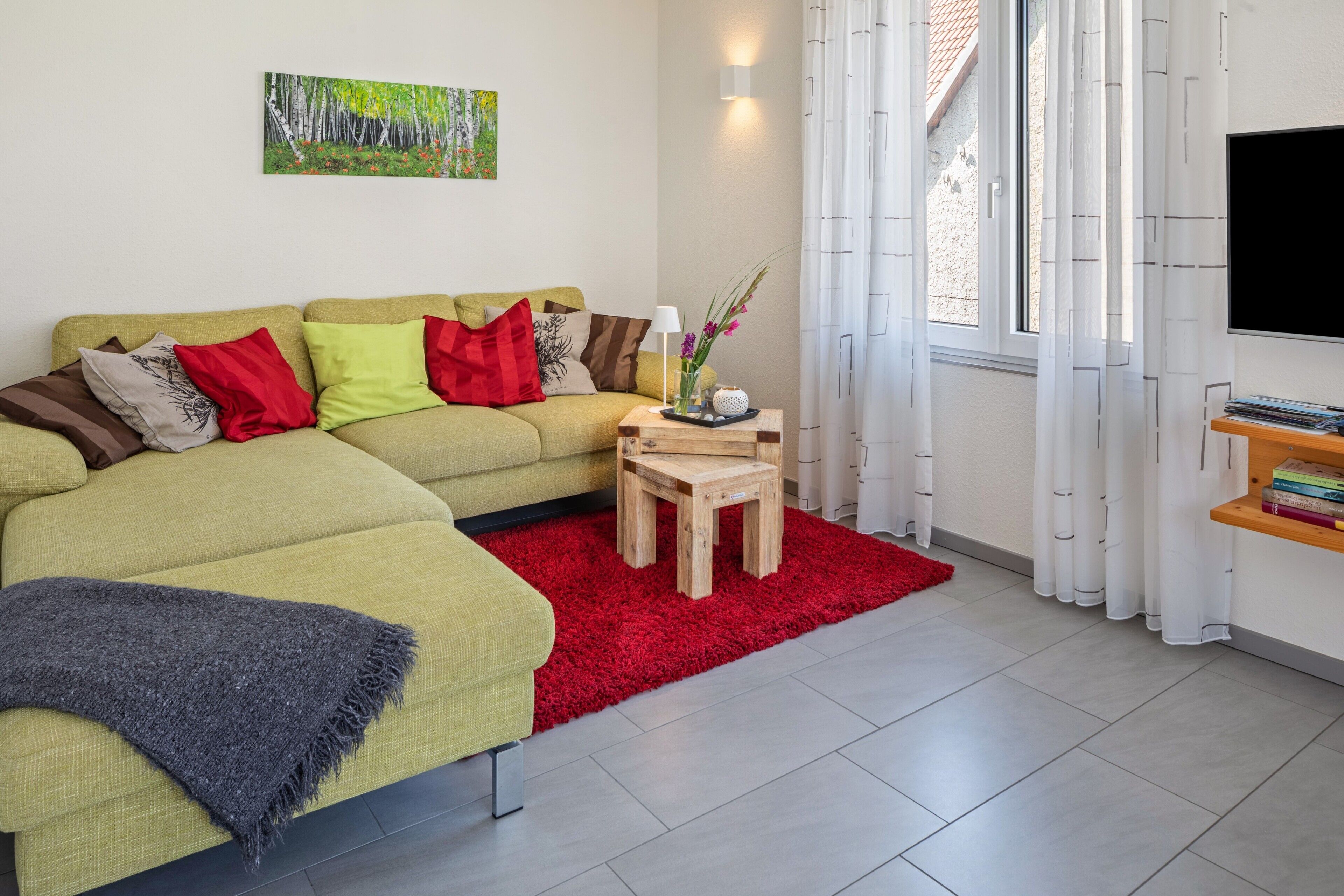 Ein modernes Wohnzimmer mit einem Ecksofa, einem kleinen Holztisch, einem roten Teppich und einem Gemälde an der Wand.