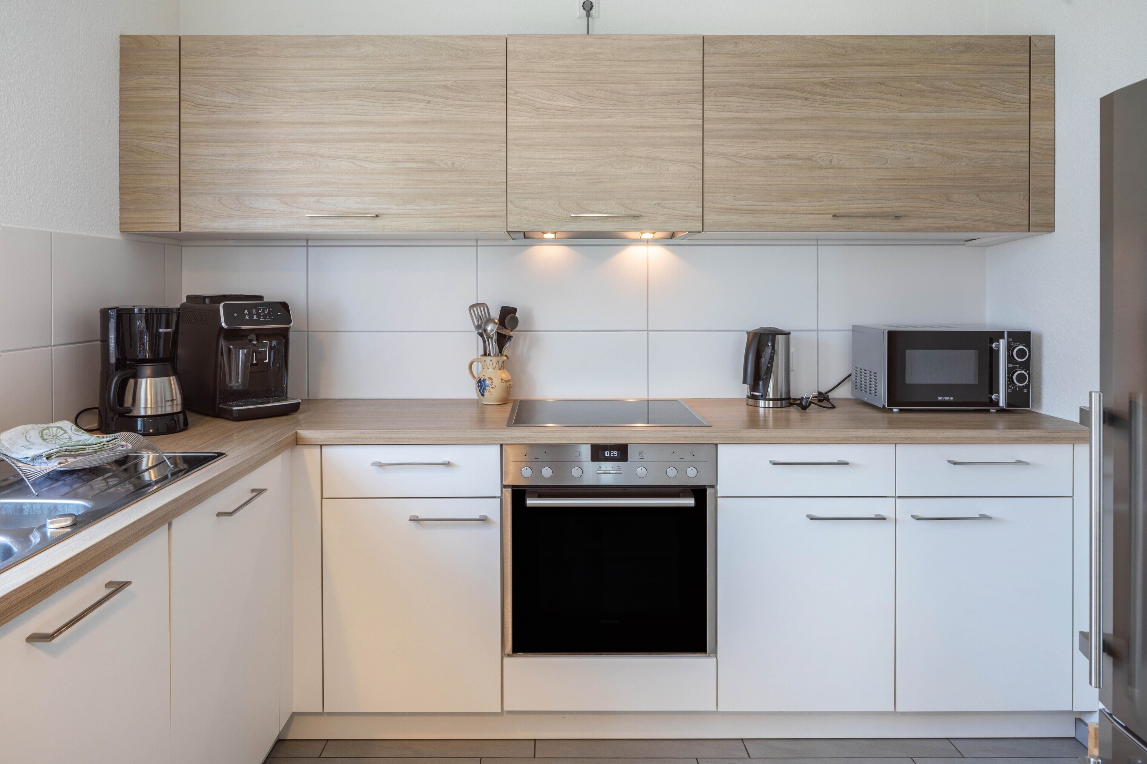 A modern kitchen with white cabinetry, a built-in oven, a microwave, a coffee maker, and a kettle.