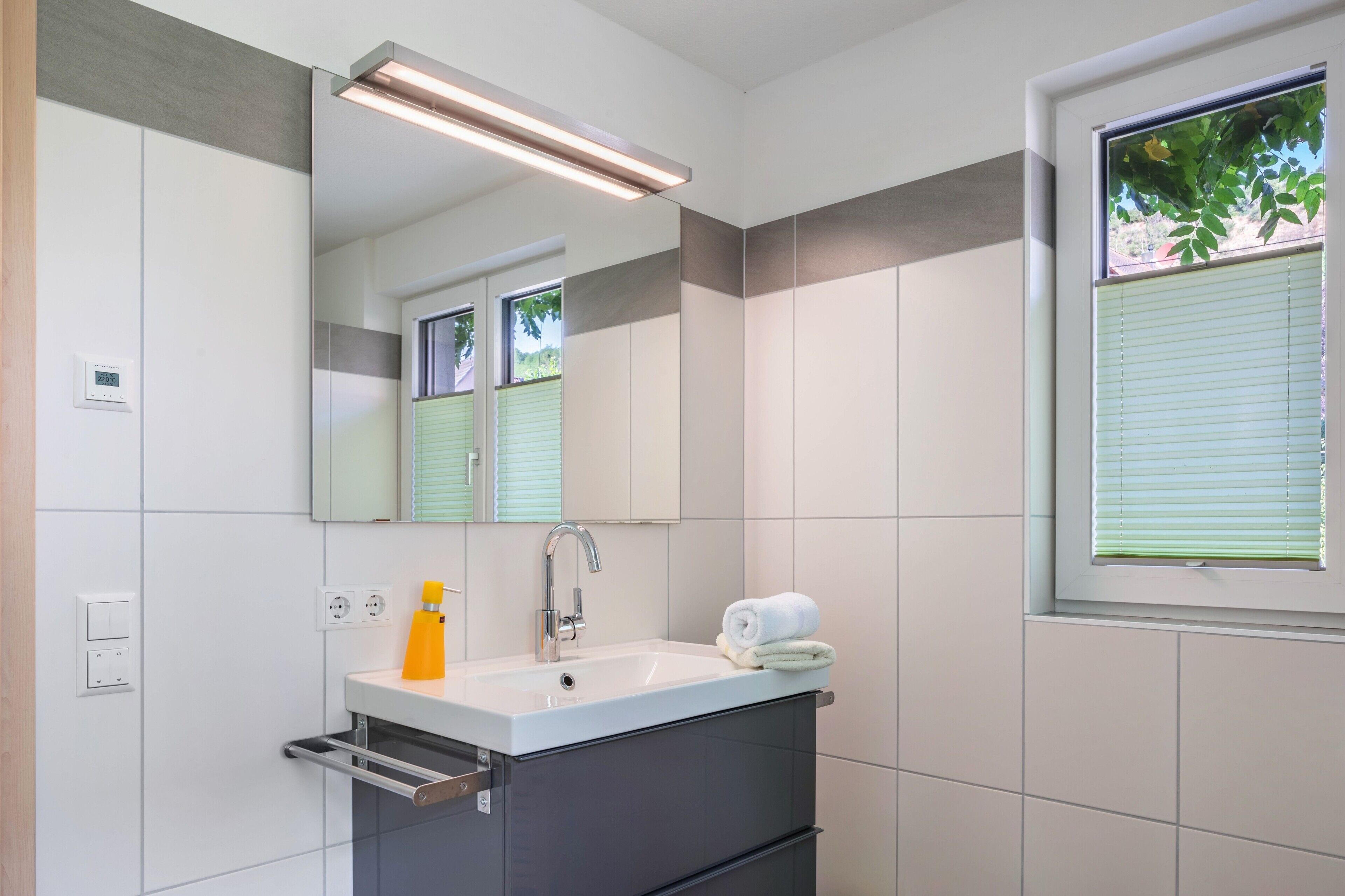 A modern bathroom with a white sink, a mirror, a towel, and a window with blinds.