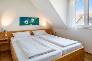 A bedroom with a wooden bed, white bedding, a painting on the wall, and a window with a view of buildings.