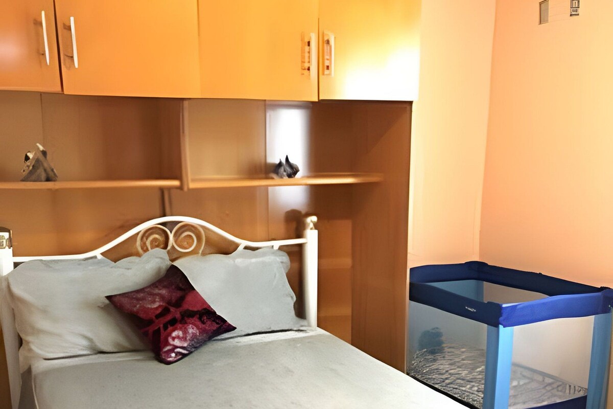Quarto com cama, travesseiro, cobertor, mesinha e uma cadeira azul.