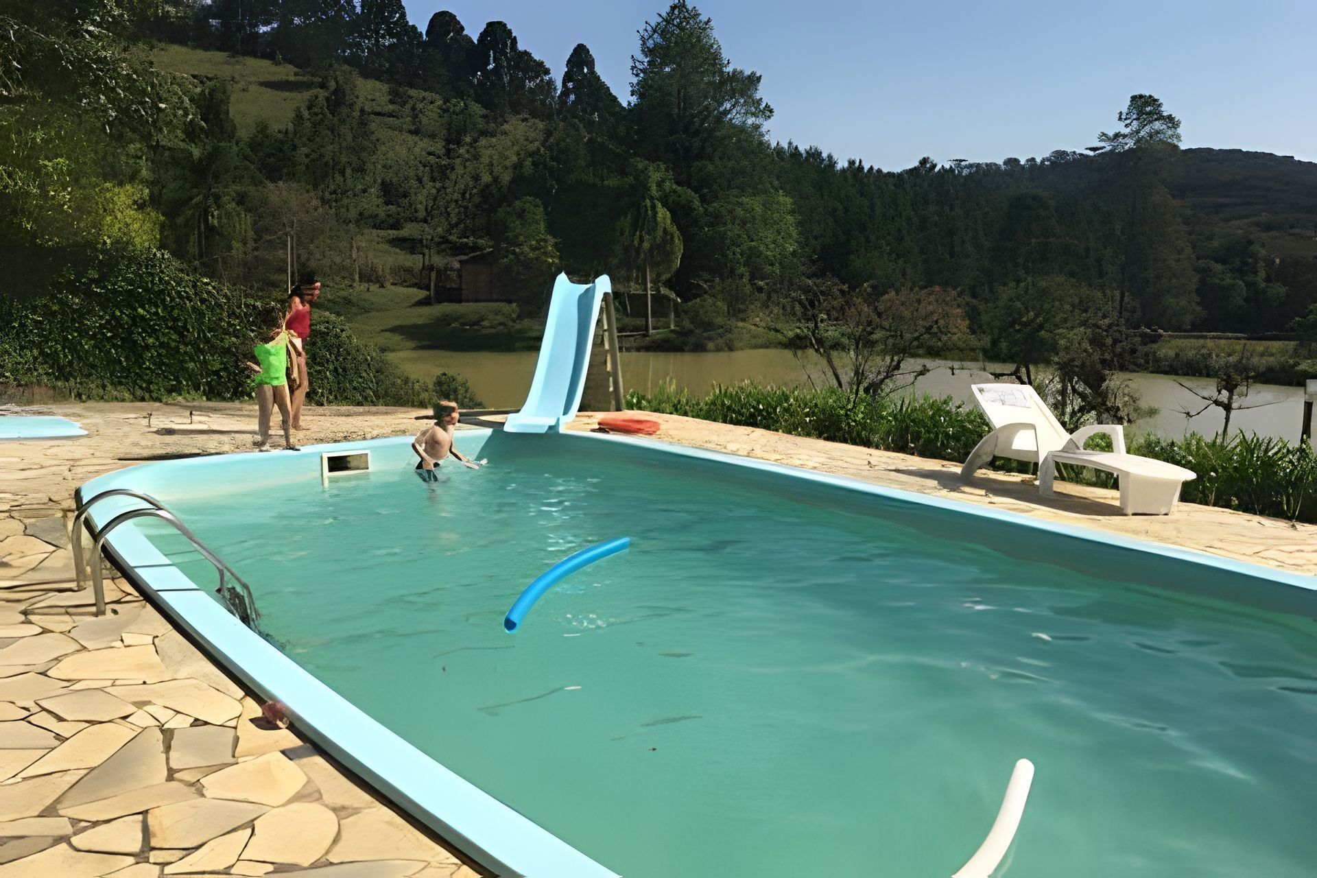 A swimming pool with a slide, surrounded by a stone deck and a view of a lake and forest.