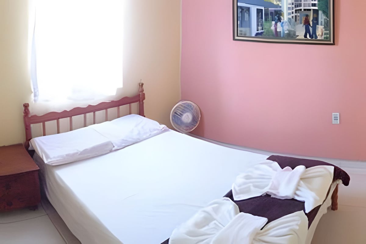 Uma cama de solteiro com lençóis brancos, cabeceira de madeira e mesa de cabeceira com superfície vermelha. Um ventilador fixado na parede é visível à direita da cama.