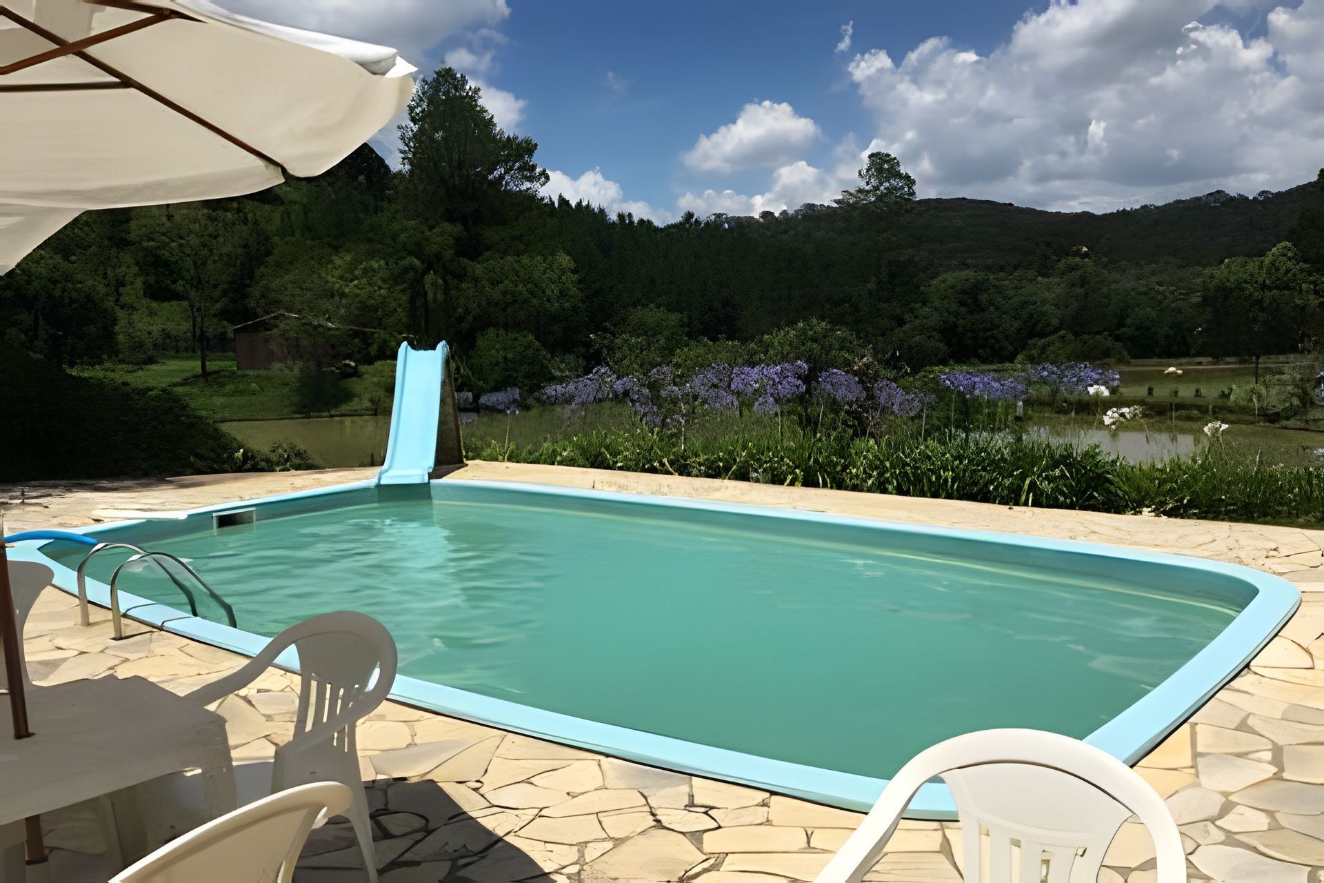 Una piscina con tobogán, rodeada de un patio de piedra, con vista a un lago y colinas al fondo.