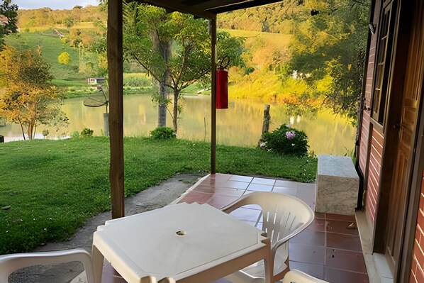 Une terrasse couverte, dotée d’une table et de chaises, qui offre une vue sur un lac et une zone verdoyante parsemée d’arbres.