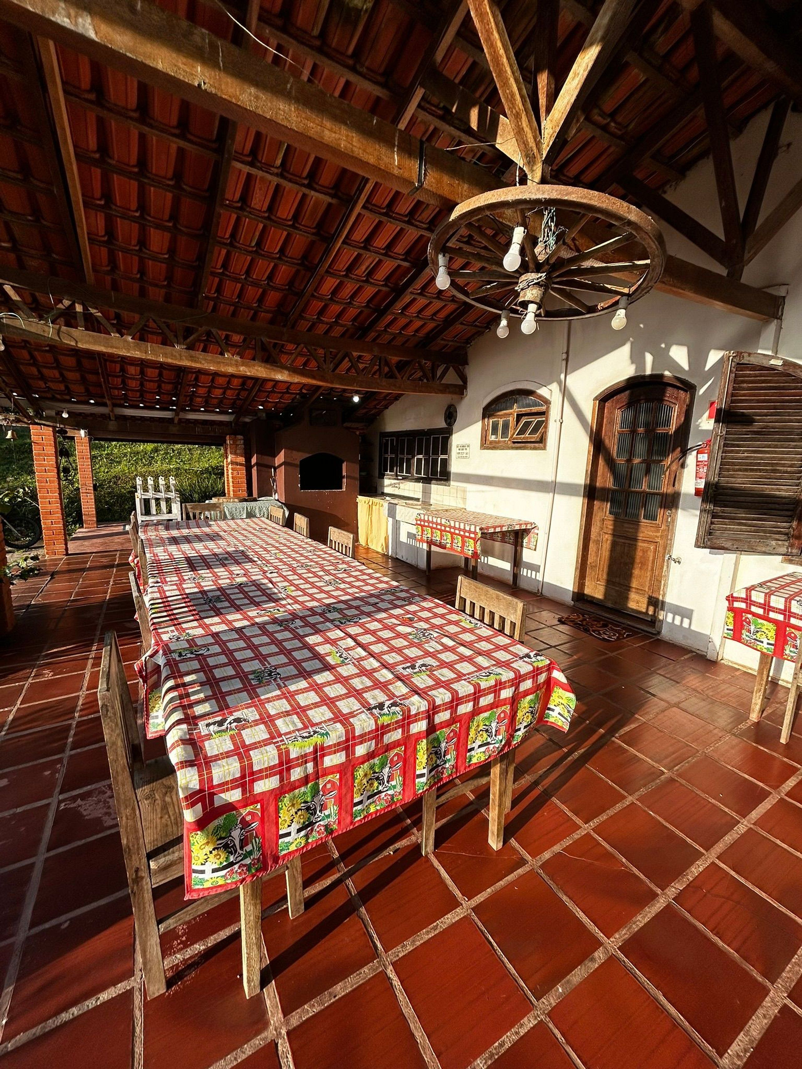 Um pátio ao ar livre com uma mesa comprida coberta por toalha xadrez, cadeiras de madeira e teto rústico com vigas aparentes.