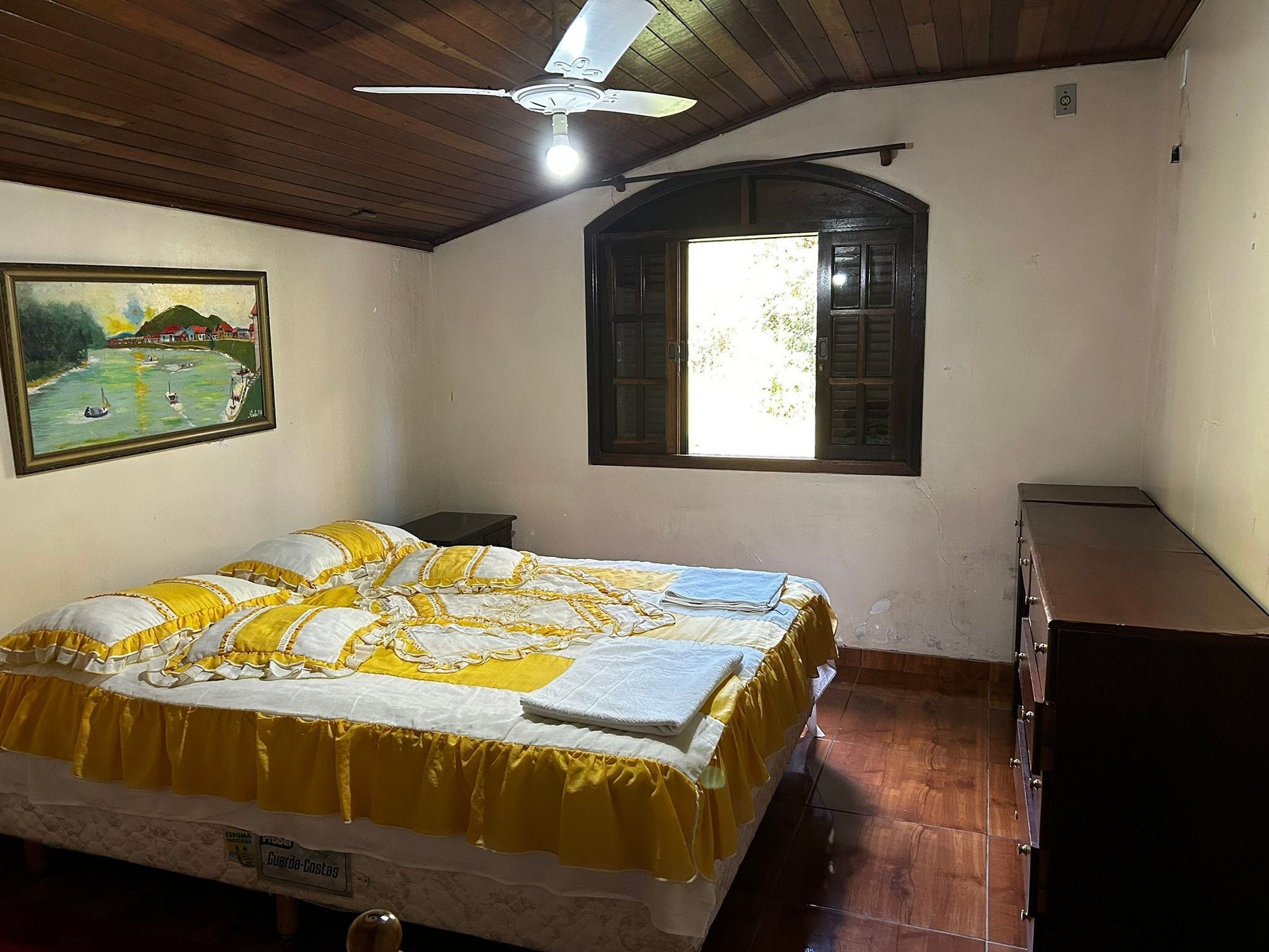 Un dormitorio con una cama, un ventilador de techo, una ventana y un cuadro en la pared.