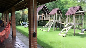 Un parque infantil con varias estructuras de juego, incluyendo un tobogán y una red de escalada, con abundante vegetación de fondo.