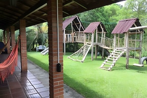 A playground with multiple play structures, including a slide and a climbing net, set against a backdrop of lush greenery.
