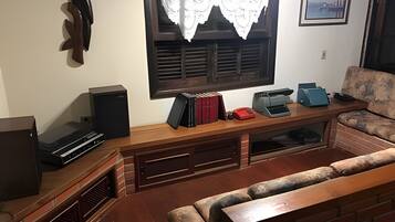 A cozy living room with a fireplace, a television, a bookshelf, and a window with a lace curtain.
