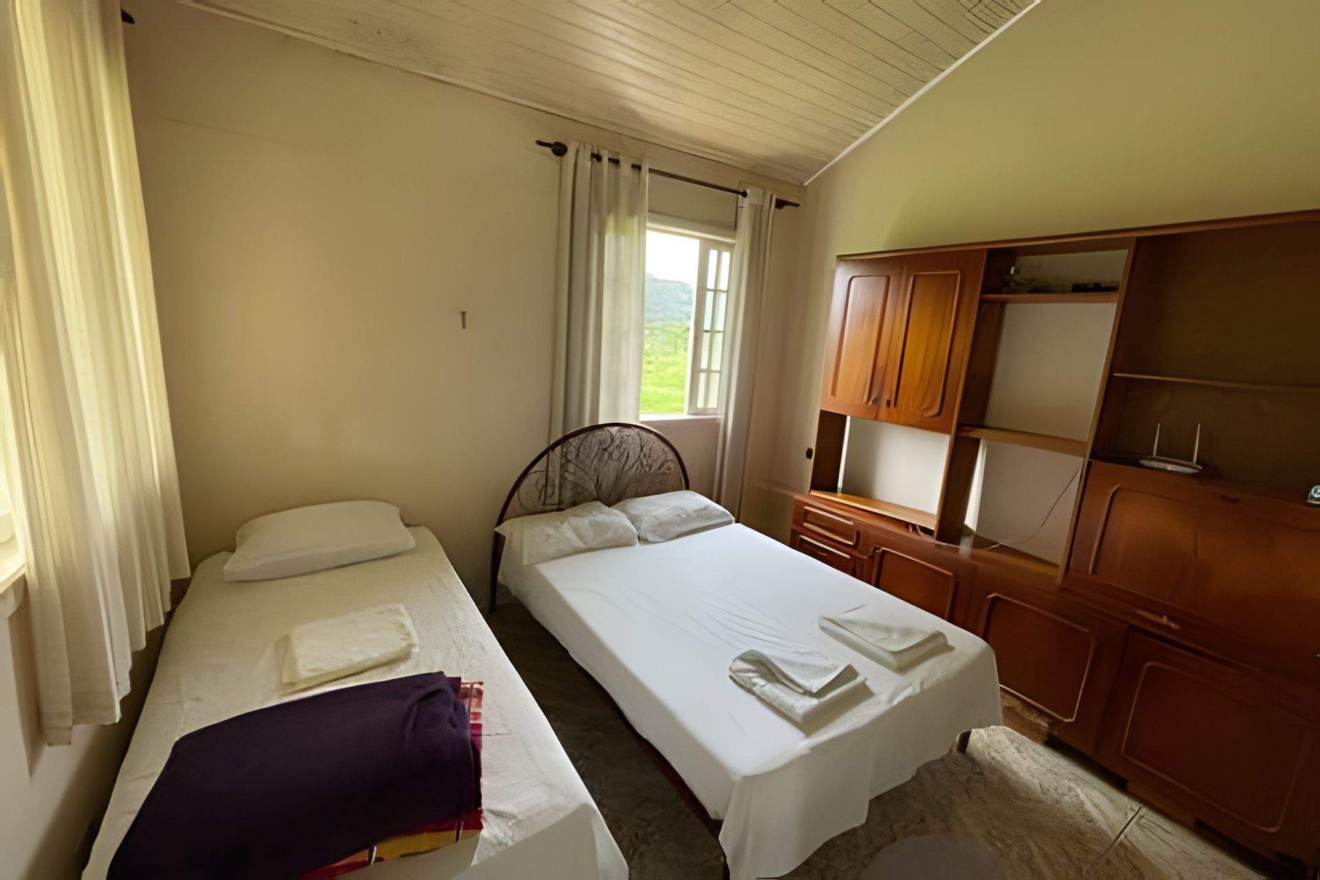 Quarto com duas camas, um guarda-roupa de madeira e uma janela com cortinas.