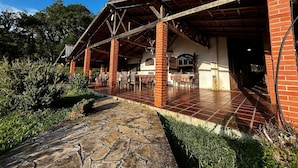 Área externa coberta com colunas de tijolo, piso de pedra e um pátio com mesas e cadeiras.