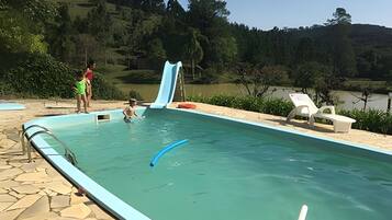 Piscina com escorrega, cercada por um deck de pedra e vista para um lago e floresta.